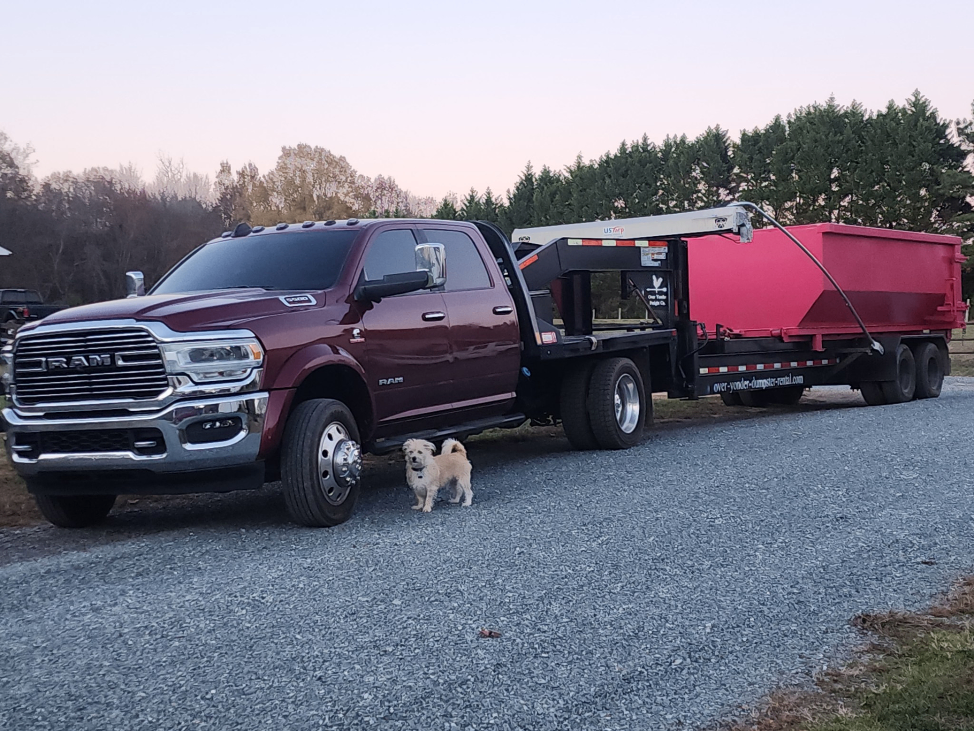 Burgundy Ram dumpster truck with red roll-off dumpster on a gravel driveway in North Carolina